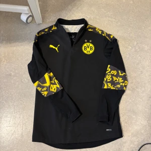 BVB långärmad träningströja Puma - Säljer en svart långärmad Borussia Dortmund träningströja från Puma med gula detaljer och grafiska tryck på axlar och ärmar. Tröjan har halv dragkedja, BVB-logga på bröstet och är tillverkad i DryCell-material som andas. Pris kan diskuteras!