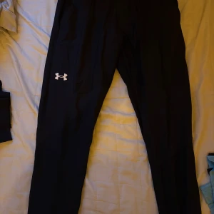 Svarta träningsbyxor från Under Armour - Svarta träningsbyxor från Under Armour med klassisk logga på låret. Byxorna har en slim passform och är gjorda i ett lätt och funktionellt syntetmaterial som passar perfekt för träning. Snygg och sportig look. Ett litet hål på framsidan som lätt kan åtgärdas