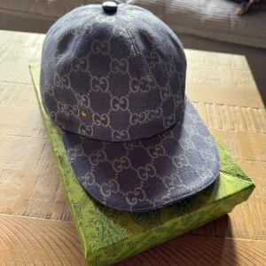 Gucci keps med GG-mönster blå - Snygg keps från Gucci i blått tyg med ikoniskt GG-mönster över hela kepsen. Justerbar rem bak i blått skinn och klassisk böjd skärm. Perfekt för dig som vill sticka ut med en lyxig accessoar. Fick den av min kusin men säljer den då jag inte tycker den passar min stil riktigt.