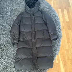Svart lång pufferjacka med huva - Säljer en svart lång pufferjacka med huva och stora fickor. Jackan har quiltad design, dragkedja och knappar framtill samt ribbade muddar vid ärmsluten. Perfekt för kalla dagar när du vill hålla dig varm och stilsäker.