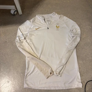 Vit Nike Dri-Fit fotbollströja Frankrike - Säljer en vit långärmad Nike Dri-Fit träningströja med Frankrikes landslagsemblem och guldfärgad swoosh. Tröjan har half zip och är i slim fit-modell, perfekt för fotbollsträning eller sportiga dagar. Pris kan diskuteras!