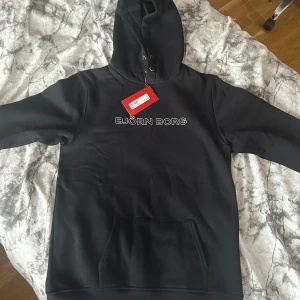 Svart hoodie från Björn Borg - Svart hoodie från Björn Borg med vit logga på bröstet. Tröjan har huva med snörning och en stor magficka framtill. Tillverkad i mjuk bomullsblandning, perfekt för chill eller till skolans kläder. Klassisk och stilren design som funkar året runt.