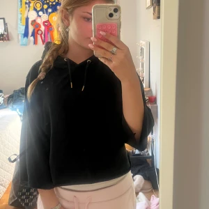 Svart croppad hoodie från 157 - Svart croppad hoodie från 157 med huva och snörning. Tröjan har vida, trekvartsärmar och är gjord i mjukt fleecematerial på insidan. Perfekt för en chill och avslappnad stil.jag ha klippt av armarna så den är trekvartsarmad och ha klippt av den så den är lite mag tröja. Passar mig som brukar ha xs s 