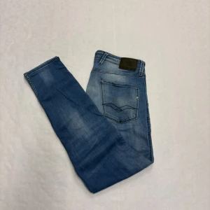 Replay Anbass - Säljer ett par ljusblå jeans från Replay med klassisk femficksdesign och raka ben. Jeansen har snygga slitningar och diskreta detaljer på bakfickorna. Perfekta för dig som gillar en avslappnad och stilren look.