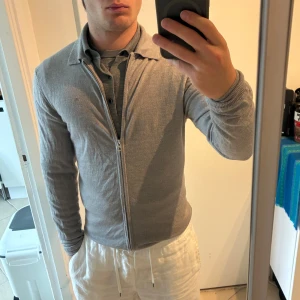 Grå kofta från J.Lindeberg med dragkedja - Grå kofta från J.Lindeberg. Plagget är i helt skick men den är tyvärr fläckig på höger bröst, vänster mage samt bak nacke. Den kommer förmodligen behöva kemtvättas, därav pris. Sitter normalt för att vara M, tröjan är uppvikt på första bilden.