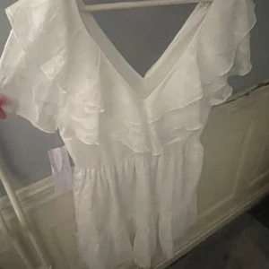 Perfekt studentklänning - Supersöt vit kortklänning med v-ringning och volanger längs axlar och bröst. Klänningen har ett lätt, transparent tyg med subtilt mönster och är kortärmad. Aldrig använd och prislapp kvar.