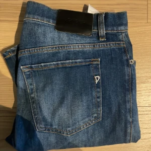 Blå jeans från Dondup, storlek 29 - Snygga blå jeans från Dondup, tillverkade i Italien. Jeansen har klassisk femficksdesign, tydliga slitningar framtill och bakficka med metallogo. De är i ett mjukt denimtyg och har normal passform med raka ben. Perfekta för dig som gillar stilrena och moderna jeans.