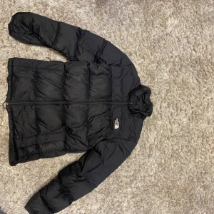 Svart pufferjacka från The North Face - Säljer en svart pufferjacka från The North Face med klassisk logga på bröstet och ryggen. Jackan har hög krage, dragkedja framtill och är quiltad för extra värme. Perfekt för kalla dagar och riktigt snygg streetstil.