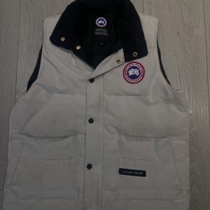 Vit dunväst från Canada Goose - Snygg vit dunväst från Canada Goose med klassisk logga på bröstet och svarta detaljer vid kragen. Västen har stora fickor framtill och stängs med både dragkedja och tryckknappar. Perfekt för lager-på-lager och riktigt chill stil.