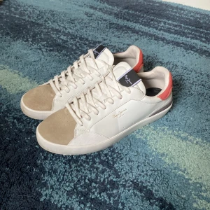 Pepe Jeans sneakers - Sneakers från Pepe Jeans i vitt skinn med beige mockadetaljer på tån och röd mocka på hälen. Klassisk låg modell med snörning och rund tå. Kommer med slitningar som nya! Nypris ligger runt 1000kr. Skriv vid minsta fundering 💭 