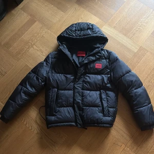 Svart pufferjacka från Hugo - Svart pufferjacka från Hugo med röd logotyp på bröstet. Jackan har huva, dragkedja framtill och två fickor med dragkedja. Den är perfekt till vintern och håller bra värme. Använd en vinter och är som ny. Pris kan diskuteras 