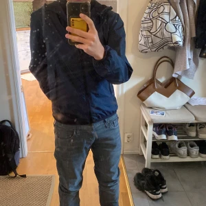 Mörkblå vindjacka med huva - Säljer en stilren mörkblå vindjacka med huva och dragkedja framtill. Jackan har en enkel design, elastiska muddar vid ärmslut och midja samt två sidofickor. Perfekt för blåsiga dagar och lätt att matcha med jeans eller sneakers.