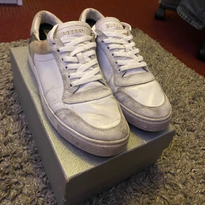 Burberry vita sneakers med rutiga detaljer - Snygga vita sneakers från Burberry med klassiska rutiga detaljer på hälen. Skorna har snörning, rund tå och är tillverkade i en mix av skinn och mocka. Perfekta för dig som vill ha en stilren men ändå lyxig look. Ikonisk Burberry-logga på plösen. Priser går att diskutera och skorna. Skick 7/10