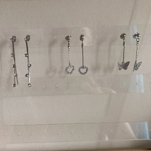 3 par silvriga hängande örhängen - Tre par hängande örhängen i silverfärg: ett par med små glittriga stenar längs smala stavar, ett par med hjärtformade hängen av strass och ett par med glittriga fjärilar. Perfekta för dig som gillar detaljer och vill ha något extra till din stil. Om du bara vill ha ett par av dem så kan du skriva till mig, då löser vi det!😽