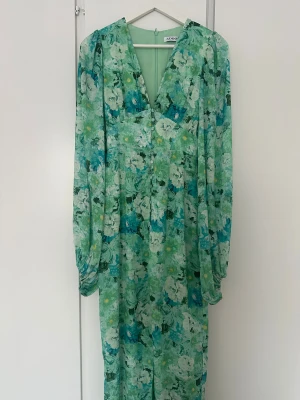 Grön blommig långklänning från Adoore - Supersnygg långklänning från Adoore i gröna och blå toner med blommigt mönster. Klänningen har v-ringning, långa ärmar och knappar framtill. Perfekt för dig som vill sticka ut med en färgglad och flowy look.