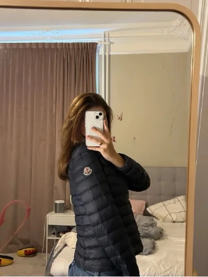 Svart dunjacka från Moncler - Säljer en svart, lätt och snygg dunjacka från Moncler med klassisk quiltad design och dragkedja framtill. Jackan har lång ärm, hög krage och ikonisk Moncler-logga på ärmen. QR FINNS, och den är garanterad äkta, storlek 14 år för den e i barn modellen, skriv ifall ni vill ha bild på qr koden. Nypris ca 7000