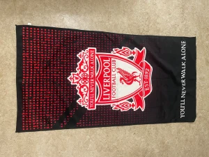 Liverpool badhandduk med klubbmärke - Stor badhandduk med Liverpool FC:s ikoniska klubbmärke i rött och vitt, samt texten 'You'll Never Walk Alone' på svart bakgrund med röda rutor. Perfekt för stranden eller poolen för dig som älskar fotboll och vill visa ditt lag.