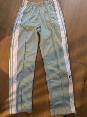 Adidas mintgröna träningsbyxor - Snygga mintgröna träningsbyxor från Adidas med klassiska vita ränder längs sidorna och tryckknappar. Byxorna har elastisk midja och sidofickor, samt Adidas logga på benet. Perfekta för chill eller sportiga dagar.