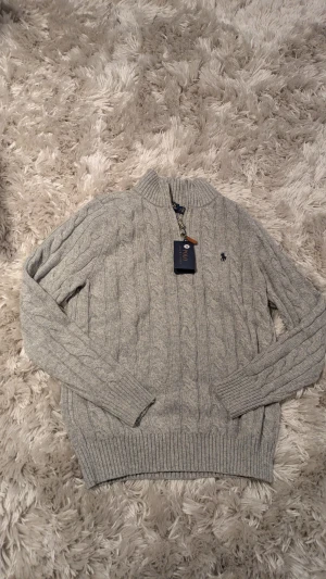 Grå stickad half zip tröja Polo Ralph Lauren - Snygg grå kabelstickad tröja från Polo Ralph Lauren med half zip och ribbad krage. Tröjan har lång ärm, klassisk broderad logga på bröstet och är gjord i mjuk ull. Perfekt för dig som gillar stilren och tidlös design.