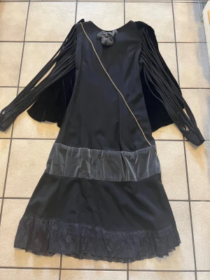 Svart långklänning med sammet och volang - Unik svart långklänning med coola öppna ärmar, sammetsdetaljer och volangkant nertill. Klänningen har en dekorativ rosett framtill, diagonal guldfärgad söm och bred sammetsmidja. Materialmix av sammet och tyg, med dragkedja bak.