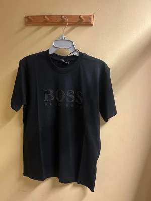 Svart t-shirt från Hugo Boss - Snygg svart t-shirt från Hugo Boss med stilrent BOSS-tryck i svart på bröstet. T-shirten har rund hals och korta ärmar, tillverkad i mjuk bomull för skön känsla. Passar perfekt till jeans eller shorts för en clean look.