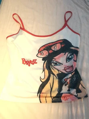 Bratz linne från SHEIN med tryck - Supersnygg vit linne-topp från SHEIN med röd kant och tunna axelband. Framsidan har ett stort Bratz-tryck med cool illustration och logga i rött. Materialet är mjuk polyester med lite stretch för skön passform. Perfekt för dig som älskar retro och statement-plagg!