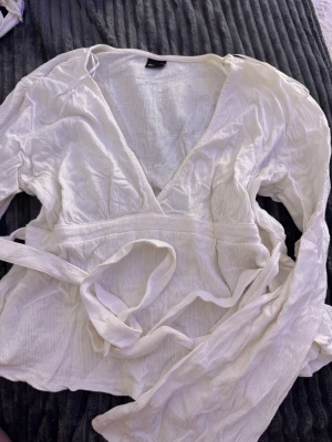 Vit omlottblus med knytband - Säljer en vit omlottblus med v-ringning och långa ärmar. Blusen har knytband i midjan och är tillverkad i ett lätt, krinklat tyg som ger en chill vibe. Perfekt för dig som gillar en luftig och stilren look.