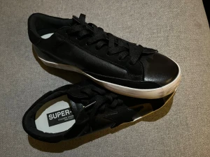 Golden Goose Super-Star svarta sneakers - Svarta Golden Goose Super-Star sneakers med vit sula och klassisk stjärndetalj på sidan. Skorna har snörning och en rund tå, tillverkade i skinn och mocka. Märkeslogga syns på hälen och innersulan. Snygg och stilren modell från Italien. Storlek 41 men funkar även på 42. Pris kan diskuteras vid snabb affär