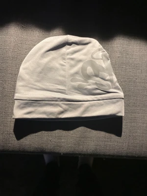 Ljusgrå mössa med tryck - Snygg ljusgrå mössa med ett coolt, ton-i-ton tryck på sidan. Mjuk och stretchig passform som sitter skönt på huvudet. Perfekt för dig som gillar stilrena accessoarer med en twist. Materialet känns som polyester och mössan har en enkel, modern design.