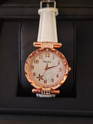 Vit klocka med roséguld och blommor - Snygg klocka från Insohic med vit rem i syntetmaterial och ett roséguldfärgat ur med glittriga stenar runt boetten. Urtavlan har blommigt mönster och tydliga siffror i guld. Perfekt accessoar för dig som gillar detaljer och stil.