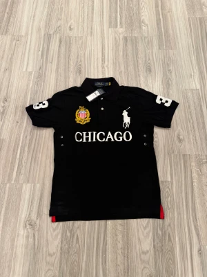 Svart polotopp från Polo Ralph Lauren - Svart polotopp med vit broderad Polo-logga och texten CHICAGO framtill. Toppens krage har en snygg röd insida, och på bröstet finns ett gult broderat emblem. Slim fit-modell med korta ärmar och vita siffror på axlarna. Perfekt för dig som gillar sportig och klassisk stil.
