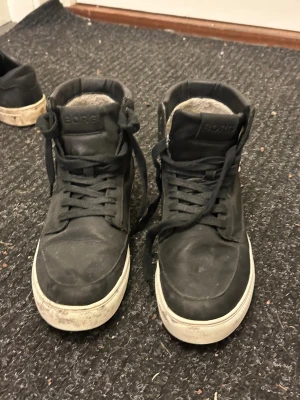 Svarta höga sneakers från Björn Borg - Säljer ett par svarta höga sneakers från Björn Borg med vit platt sula och snörning. Skorna har en rund tå och är tillverkade i skinn med vadderad kant runt ankeln för extra komfort. Klassisk stil med diskret logga på plösen och hälen.