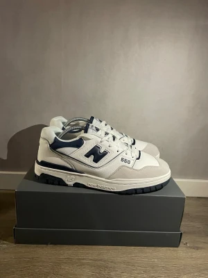 New Balance 550  marinblå  - Säljer ett par New Balance 550 sneakers i vitt och marinblått. Skorna har klassisk retrodesign med läder och mockadetaljer, tydlig NB-logga på hälen och sidan samt snörning framtill. Snygg platt sula och perforerade paneler för extra stil.