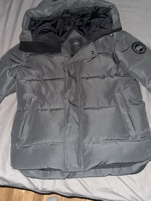 Mörkgrå pufferjacka från Canada Goose black label - Säljer en mörkgrå pufferjacka från Canada Goose, modell macmillan. Jackan har huva, broderad logga på ärmen och praktiska fickor. Materialet är polyester och dun, perfekt för kalla dagar. Stängs med dragkedja och tryckknappar. Snygg och funktionell design.