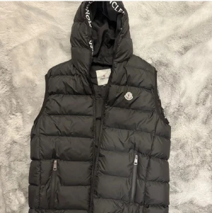 Svart dunväst från Moncler med huva - Svart dunväst från Moncler med stor huva och logotyp på bröstet. Västen har quiltad design, två fickor med dragkedja och Moncler-tryck längs huvan. Perfekt för lager-på-lager och streetstyle. Stängs med dragkedja framtill.
