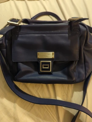 Blå axelväska Weekend Max Mara - Snygg blå axelväska från Weekend Max Mara i mjukt skinn med guldfärgade metalldetaljer och justerbar axelrem. Väskan har ett stilrent lock med dekorativt spänne och insidan är fodrad med beige tyg och svarta prickar. Perfekt för dig som gillar klassisk design med en twist.