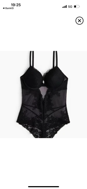 Svart spetsig body med meshdetaljer - Svart body med tunna axelband, vadderade kupor och genomskinliga meshpartier. Spetsdetaljer längs kanterna och över hela bodyn ger en lyxig och edgy vibe. Perfekt för dig som gillar att sticka ut med din stil.