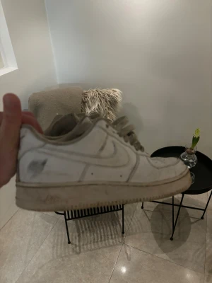 Nike Air Force 1 vita sneakers - Klassiska Nike Air Force 1 sneakers i vitt med låg profil och rund tå. Skorna har snörning och är tillverkade i skinn med en platt sula. Perfekt för dig som gillar streetwear och vill ha en ikonisk sneaker i garderoben.
