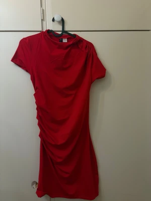 Röd kort klänning från H&M - Snygg röd kort klänning från H&M med draperad detalj på sidan. Klänningen har rund halsringning och korta ärmar, perfekt för dig som vill sticka ut med en enkel men cool look. Tillverkad i mjukt bomullsmaterial som känns skönt mot huden.