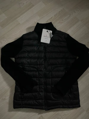 Svart dunväst från Moncler M - Svart dunväst från Moncler med quiltad front, dragkedja och ribbstickade ärmar. Klassisk hög krage och ikonisk Moncler-logga på bröstet. Västen har en clean look och är perfekt för lager-på-lager. Snygg och tidlös design som funkar året runt.