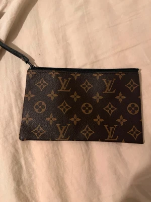 brun kuvertväska för smink - Snygg brun kuvertväska med klassiskt monogrammönster i guld. Väskan har en praktisk dragkedja och en handledsrem i svart. Perfekt storlek för att bära med sig det viktigaste och ger en lyxig touch till din outfit.