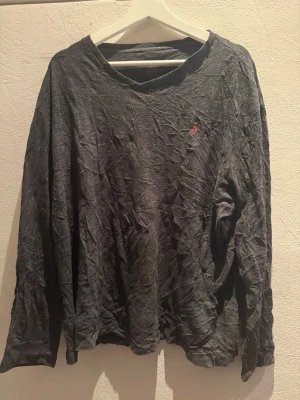 Mörkgrå långärmad tröja från Polo Ralph Lauren - Mörkgrå långärmad tröja från Polo Ralph Lauren med klassisk röd broderad logga på bröstet. Tröjan har en rund halsringning och är gjord i mjuk bomull. Perfekt för chill dagar och enkel att matcha med jeans eller joggers.                                                          OBS! Polo loggan är utsliten på insidan då de blev fel i tvättningen! 