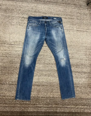 Replay Waitom blå jeans, storlek 32 - Snygga Replay Waitom jeans i klassisk blå tvätt med slitningar och ett coolt hål på ena benet. Jeansen har raka ben, fem fickor och läderpatch bak i midjan. Perfekta för dig som gillar en avslappnad och trendig look.
