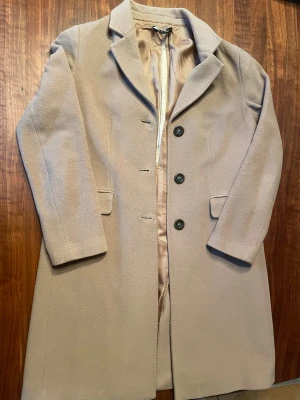 Beige ullkappa från Miroglio Fashion - Stilren beige kappa i ull med klassisk krage och tre svarta knappar framtill. Fodret är ljusbeige och jackan har två fickor med lock. Perfekt för dig som gillar en clean och tidlös look.