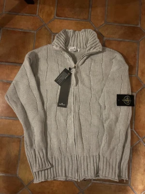 Ljusgrå stickad kofta från Stone Island - Snygg ljusgrå stickad kofta från Stone Island med ribbad krage och muddar. Koftan har dragkedja framtill och det klassiska Stone Island-märket på ärmen. Diskret mönster i stickningen ger extra vibe. Perfekt för dig som gillar clean och stilren look.