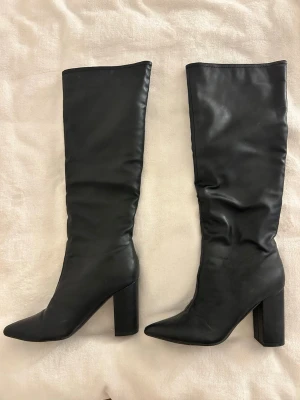 Svarta höga stövlar med klack - Säljer ett par svarta höga stövlar med spetsig tå och rejäl blockklack. Stövlarna är tillverkade i skinnimitation och har en stilren, minimalistisk look som passar till många outfits. Perfekt för dig som gillar att sticka ut med snygga boots.