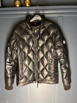 Svart quiltad dunjacka från Moncler - Svart dunjacka från Moncler med quiltat mönster och ribbade muddar. Jackan har hög krage, två fickor med dragkedja och Moncler-logga på ärmen. Materialet är glansig polyester och dunfyllning. Perfekt för kalla dagar och har en clean, modern look.