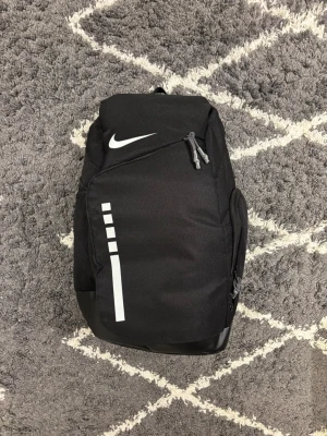 Nike Elite bag - Snygg svart ryggsäck från Nike med vita detaljer och logga. Rymligt huvudfack, flera dragkedjefickor och vadderade axelremmar med coola transparenta partier. Perfekt för skolan, gymmet eller vardagsäventyr. 
