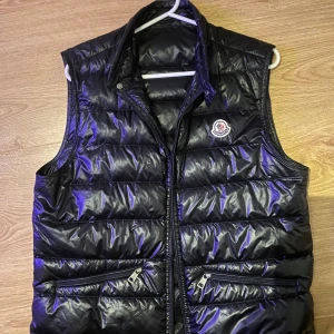 Svart dunväst från Moncler - Äkta Svart, glansig dunväst från Moncler med quiltad design och ståkrage. Västen har två framfickor med dragkedja och Moncler-logga på bröstet. Perfekt för lager-på-lager och har en stilren look som funkar till många outfits. Drag kedjan har gott sönder så den är borta därav så vet jag inte exakt villket pris jag ska lägga så KOM MED BUD!