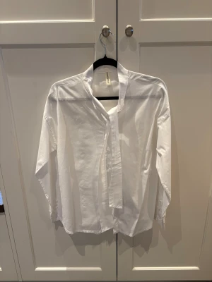 Vit knytblus - Vit knytblus mes ståkrage och knappar framtill. Skjortan har lång ärm och är tillverkad i mjuk bomull som känns skön mot huden. Perfekt för dig som gillar stilrena och enkla plagg med modern touch. Inköpt på Two Angels i Göteborg 
